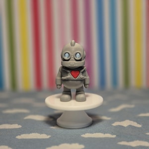May include: A grey robot figurine with a red heart on its chest stands on a small white pedestal. The robot has large, round eyes and a detailed design. The background features a colourful striped pattern.