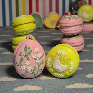 May include: A selection of colourful, decorative macarons and egg-shaped ornaments. The macarons are pink and yellow, with white filling. The ornaments feature floral and chicken designs. The background has a striped pattern.