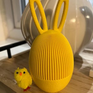Puede incluir: Una decoración de Pascua amarilla brillante con forma de conejito, con detalles de costillas verticales. El conejito tiene orejas altas y erguidas. Un pollito amarillo pequeño y esponjoso con patas rojas y pico naranja está cerca.