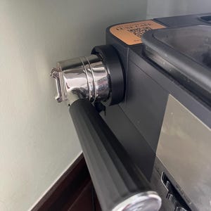 May include: A black espresso machine with a chrome portafilter inserted into the group head. The machine features a black handle with a silver end cap and a tan sticker on top with the text LUXE CAFE and a QR code. A grind size dial is visible.