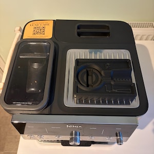 May include: A black and silver Ninja Luxe Cafe coffee machine. The appliance has a sleek design with a silver control panel and two knobs. The top features a black lid and a yellow sticker with the text "LUXE CAFE" and QR codes. The machine is designed to use whole beans only.