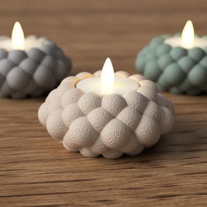 May include: Three decorative candle holders with a textured, bubble-like design. The holders are in shades of grey, white, and green, each holding a lit candle. The candles emit a warm glow, creating a cosy ambiance. The holders are placed on a wooden surface.