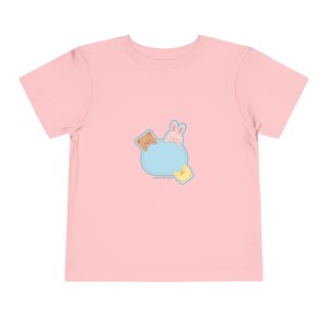 May include: A light pink toddler t-shirt featuring a cartoon design. The design includes a blue oval with a brown bear, pink bunny, and yellow bear. The text "just a little part" is printed below the oval.