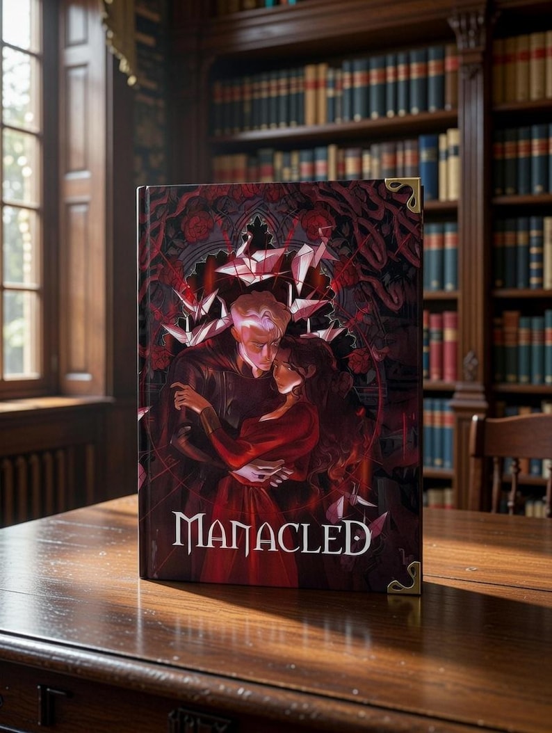 May include: A hardback book titled "Manacled" with a dark red and black cover depicting a couple embracing. The cover art includes gothic architectural elements and floral designs. The book has gold corner protectors and is on a wooden table.