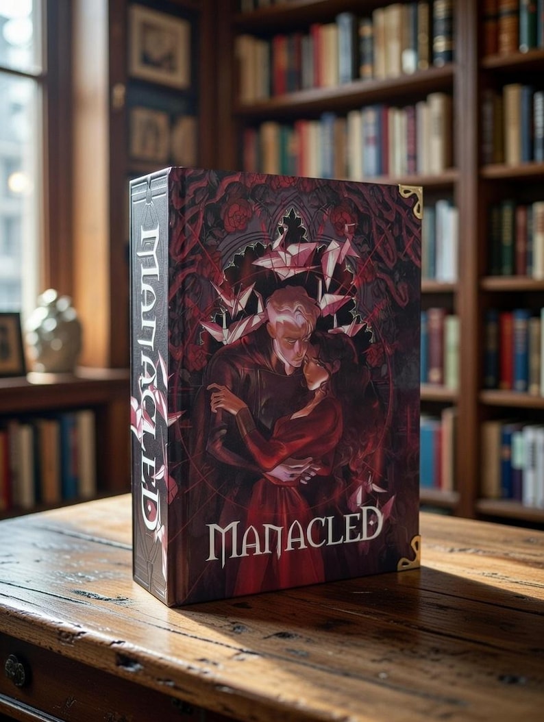 May include: A hardback book titled "Manacled" is displayed on a wooden table. The cover showcases a dark, romantic illustration in red and black, with two figures embracing. The spine shows the title vertically. A library forms the backdrop.