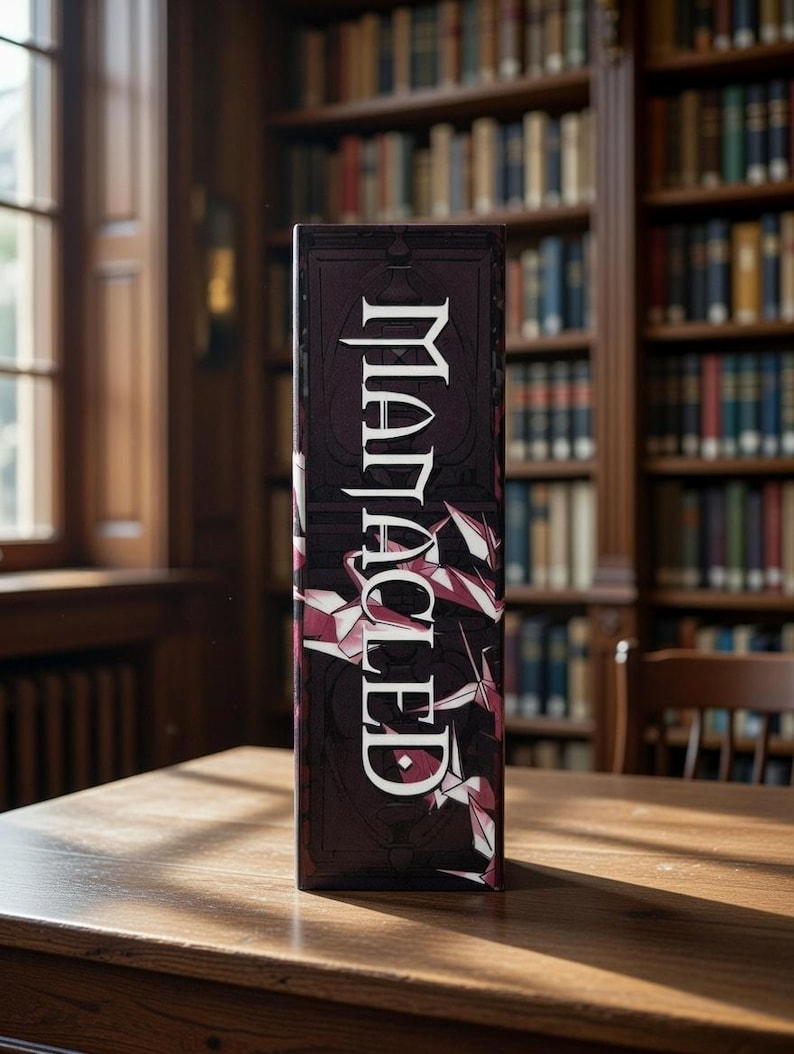 May include: A rectangular box with the word "Malacled" in white, stylized lettering. The box has a dark burgundy background with geometric shapes in white and pink. The box is on a wooden table in front of a bookshelf.