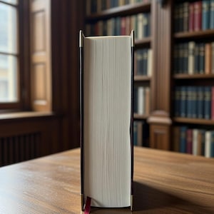 May include: A large hardback book with cream-coloured pages, a black spine, and gold-coloured metal corners. A red ribbon bookmark is inserted. The book is on a wooden table in front of a bookshelf.