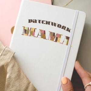 Könnte beinhalten: Ein weißes Notizbuch mit dem Text "PATCHWORK MAMA" auf dem Cover. Das Wort "MAMA" ist mit einem Patchwork-Design in Braun-, Rosa-, Gelb- und Grüntönen versehen. Ein weißes Gummiband ist sichtbar.