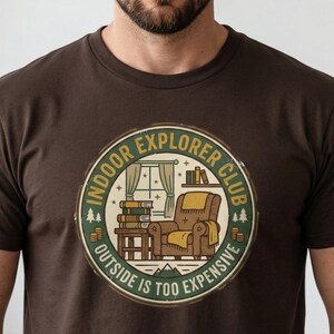 Può includere: T-shirt marrone con grafica circolare. La grafica presenta una scena interna accogliente con una poltrona, libri e una finestra, con il testo "INDOOR EXPLORER CLUB" e "OUTSIDE IS TOO EXPENSIVE".
