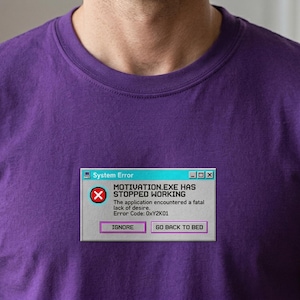 Könnte beinhalten: Lilafarbenes T-Shirt mit einem humorvollen "System Error"-Grafik. Die Grafik imitiert eine Computerfehlermeldung mit der Aufschrift "MOTIVATION.EXE HAS STOPPED WORKING" und den Optionen "IGNORE" oder "GO BACK TO BED".