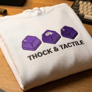 May include: White hoodie featuring embroidered purple keycaps and the text "THOCK & TACTILE". The keycaps have various designs, including a lotus flower. The hoodie is folded on a wooden surface.
