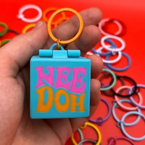 Könnte beinhalten: Ein blauer quadratischer Schlüsselanhänger mit dem Wort "NEE DOH" in rosa und orangefarbenen Buchstaben. Der Schlüsselanhänger hat einen kleinen orangefarbenen Ring. Andere bunte Ringe sind auf rotem Hintergrund verstreut.