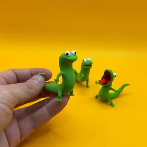 May include: Three green lizard figurines with white eyes and open mouths are displayed against a yellow background. One lizard is held by a hand. The lizards have a cartoonish design and are in various poses.