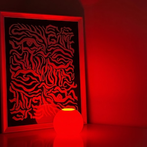 Może przedstawiać: Czerwona, kulista lampa emituje delikatną poświatę, umieszczona obok oprawionego dzieła sztuki. Grafika przedstawia czarne tło z czerwonymi, abstrakcyjnymi liniami. Pokój jest oświetlony czerwonym światłem.