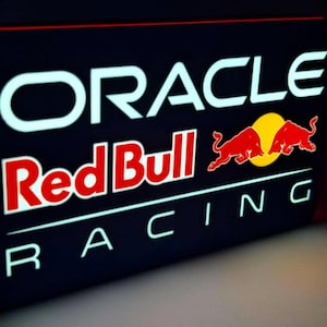 May include: A rectangular sign featuring the words "ORACLE" and "Red Bull RACING" in white and red. The Red Bull logo, with two red bulls and a yellow sun, is also visible. The sign has a black background and a red border.