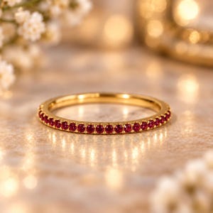 Puede incluir: Un anillo de banda fina de oro que presenta una hilera continua de pequeños rubíes rojos redondos y vibrantes engastados a lo largo de toda la circunferencia. El metal tiene un acabado pulido y las piedras son de tamaño uniforme.
