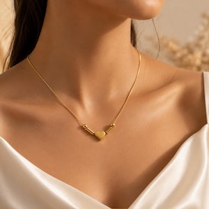 May include: A gold-toned necklace featuring a central heart-shaped charm flanked by two small cylindrical beads and two spherical beads on a thin chain. The metal has a smooth, polished finish, creating a consistent gold appearance throughout the piece.