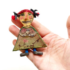 May include: A small figurine crafted from paper and metal, featuring a figure with red braided hair, black bows, and a triangular green dress decorated with floral patterns and a silver-toned Claddagh emblem at the center of the chest.