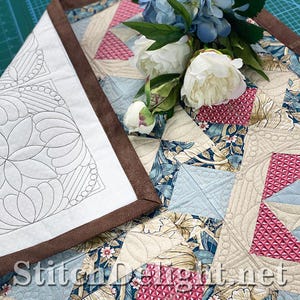 Può includere: Un runner da tavola trapuntato con un design patchwork in motivi floreali e geometrici blu, crema e rosa, bordato da tessuto marrone. Il pezzo presenta dettagli di cucitura complessi e il testo Stitch Delight net.