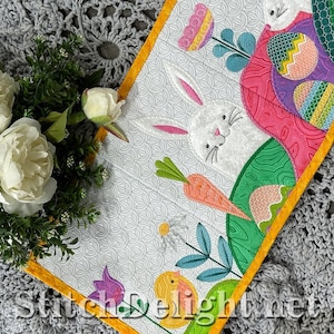 May include: A white Easter-themed table runner with a bunny, carrots, and Easter eggs. The runner has a yellow border and the text "StitchDelight.net" at the bottom. The background is a gray crocheted pattern.