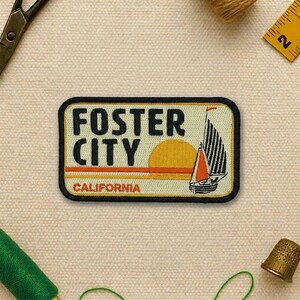 May include: Embroidered patch featuring "FOSTER CITY CALIFORNIA" in black and orange text. The design includes a sailboat with black and white sails, a setting sun, and a black border. The patch is rectangular with rounded corners.