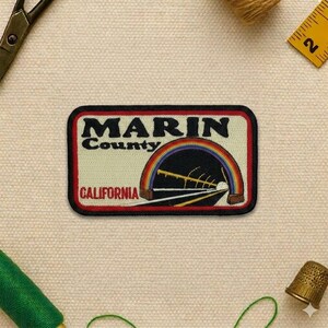 May include: A rectangular patch with a black border features the words "MARIN County CALIFORNIA" in black and red. A rainbow arches over a tunnel and road design. The patch is on a beige fabric background.