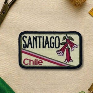May include: Embroidered patch featuring the word "SANTIAGO" above "Chile" in red text. The patch has a cream background with a diagonal red and pink stripe and a floral illustration. The patch is rectangular with rounded corners and a dark green border.