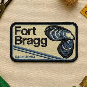 May include: Rectangular embroidered patch featuring the words "Fort Bragg" in black, with a blue and white mussel shell design. The word "CALIFORNIA" is below the text. The patch has a black border and a light beige background.