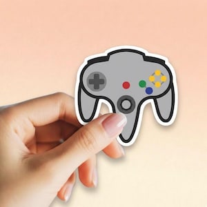 May include: A gray sticker of a classic video game controller with a black outline. The controller has a directional pad, red, green, blue, and yellow buttons, and a central button. The sticker is held by a hand.