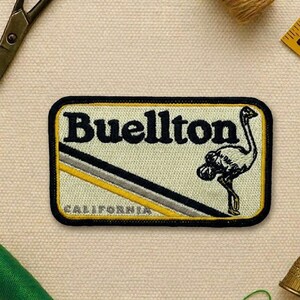 May include: Embroidered patch with the word "Buellton" in black, featuring an ostrich illustration. The patch has a black border, with yellow and gray diagonal stripes. The word "CALIFORNIA" is at the bottom.