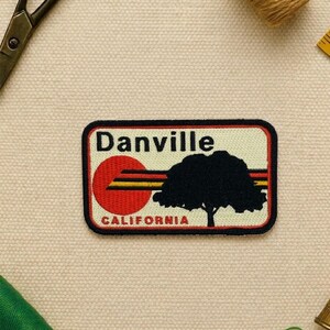 May include: A rectangular patch with a black border features the word "Danville" above a red sun design with yellow and black stripes. A black tree silhouette is on the right, with "CALIFORNIA" below.