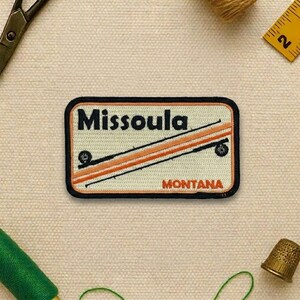 May include: Rectangular patch with the word "Missoula" in black letters above a design of orange and black lines. The word "MONTANA" is in orange letters below the design. The patch has a black border.