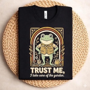 May include: Black t-shirt featuring a cartoon frog in a vest and bow tie, standing in a garden setting. The design includes a rainbow arch and the text "TRUST ME, I take care of the garden."