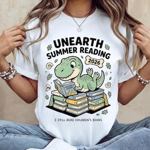 May include: White t-shirt with a green dinosaur reading a book titled "Children's Story." The shirt features the text "UNEARTH SUMMER READING 2026" and "I STILL READ CHILDREN'S BOOKS."