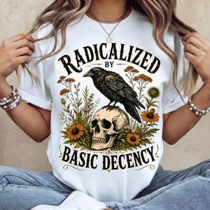 Könnte beinhalten: Ein weißes Kurzarm-T-Shirt mit einem Grafikdruck eines schwarzen Raben, der auf einem menschlichen Schädel sitzt, umgeben von orangefarbenen und gelben Wildblumen. Der Text RADICALIZED BY BASIC DECENCY ist in schwarzen Buchstaben gedruckt.