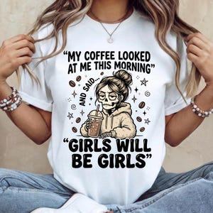 May include: White t-shirt with a graphic featuring a skeleton-faced woman holding a coffee drink. The text reads "MY COFFEE LOOKED AT ME THIS MORNING AND SAID... GIRLS WILL BE GIRLS" with coffee bean and star illustrations.