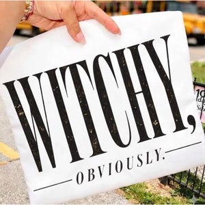 May include: A white rectangular fabric item featuring the black text WITCHY, in a large distressed serif font, followed by the word OBVIOUSLY, in a smaller serif font, underlined by a horizontal line on both sides of the word.