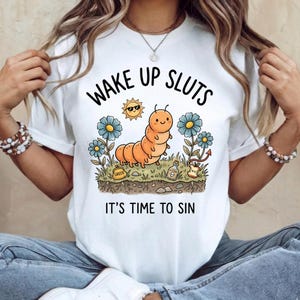 May include: White t-shirt with the text "WAKE UP SLUTS" above an illustration of an orange caterpillar, a smiling sun, blue flowers, and the phrase "IT'S TIME TO SIN". The graphic design includes elements of nature and a playful aesthetic.