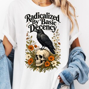 Könnte beinhalten: Ein weißes T-Shirt mit einem Grafikdruck eines schwarzen Raben, der auf einem menschlichen Schädel sitzt, umgeben von orangefarbenen und weißen Blumen. Der Text "Radicalized By Basic Decency" ist in einer fetten, schwarzen Schriftart gedruckt.
