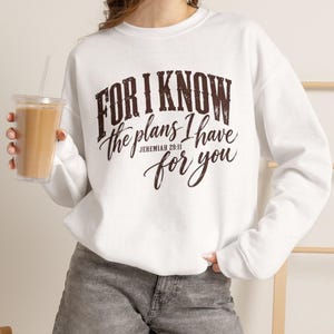 May include: A white crewneck sweatshirt featuring dark brown text that reads, "FOR I KNOW the plans I have for you JEREMIAH 29:11". The sweatshirt has long sleeves and a ribbed collar, cuffs, and hem, providing a clean and simple look for casual wear.