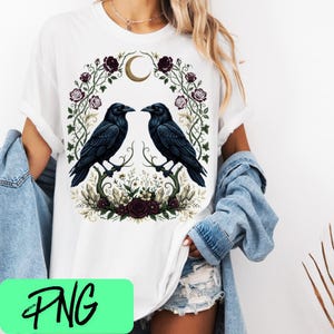 May include: A white short-sleeved t-shirt featuring a graphic print of two black ravens facing each other, surrounded by a wreath of dark red roses, green vines, and a golden crescent moon, with crystals and flowers at the base of the design.