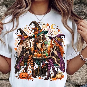 May include: A white t-shirt featuring a colourful graphic of three skeleton witches wearing pointed hats, sitting at a table with pumpkins, ghosts, and a black cat. The witches have long hair and are dressed in capes, striped stockings, and boots.