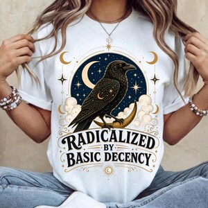 Pode incluir: Uma t-shirt branca com um gráfico de um corvo preto pousado numa lua crescente dourada. O design inclui estrelas, nuvens e o texto "RADICALIZED BY BASIC DECENCY" numa fonte escura. A ilustração está dentro de uma moldura circular.