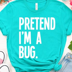 Può includere: Una maglietta turchese con la scritta bianca "PRETEND I'M A BUG". La maglietta ha le maniche arrotolate e un nodo sull'orlo. L'immagine mostra anche occhiali da sole, scarpe rosa e pantaloncini di jeans.