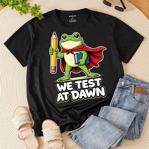 Op de afbeelding: Zwart T-shirt met een cartoon kikker superheld die een potlood en een boek vasthoudt. De kikker draagt een rode cape en de tekst "WE TEST AT DAWN" staat eronder. Het shirt wordt gecombineerd met een blauwe jeans en sandalen.