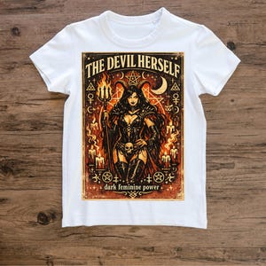 Puede incluir: Camiseta blanca con un gráfico de una diablesa sosteniendo un tridente, rodeada de velas y símbolos ocultos. El texto en la camiseta dice "THE DEVIL HERSELF" y "dark feminine power". El diseño es en tonos marrones, negros y naranjas.
