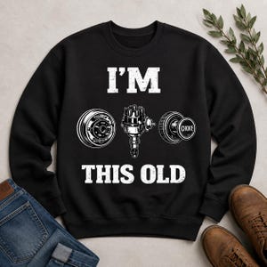 Puede incluir: Sudadera negra con el texto "I'M THIS OLD" e ilustraciones blancas de piezas de automóviles: una rueda, un distribuidor y un estrangulador. La sudadera se combina con vaqueros azules y botas de cuero marrones.