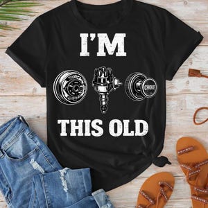Puede incluir: Camiseta negra con el texto "I'M THIS OLD" e imágenes de piezas de automóviles. La camiseta presenta un tambor de freno redondo, un distribuidor y un estrangulador. La camiseta se combina con vaqueros azules y sandalias marrones.