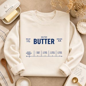 Könnte beinhalten: Cremefarbenes Sweatshirt mit dem Wort "BUTTER" in Marineblau und Umrechnungen für Backmaße. Der Text enthält "SALTED", "GRADE AA" und "NET WT. 4 OZ. (113g)." Eine Butterdose und Backzutaten sind im Hintergrund zu sehen.