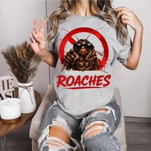 May include: A light gray t-shirt featuring a graphic of a cockroach wearing sunglasses, inside a red circle with a line through it. The text "BUENAS NOCHES ROACHES" is printed below the graphic in red.